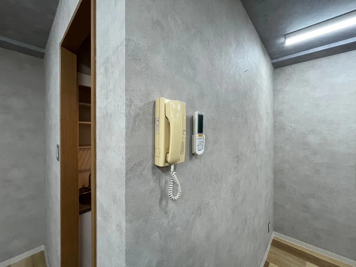- グレーの壁に設置された電話機とインターホンのある部屋、入り口が見えるシンプルな内装